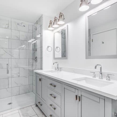 home improvement contractors A newly-remodeled home bathroom featuring a double-vanity sink, white tile floors and shower walls, and marble counters.
