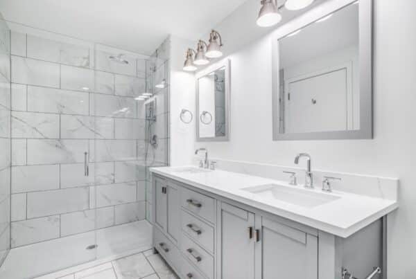 A newly-remodeled home bathroom featuring a double-vanity sink, white tile floors and shower walls, and marble counters.