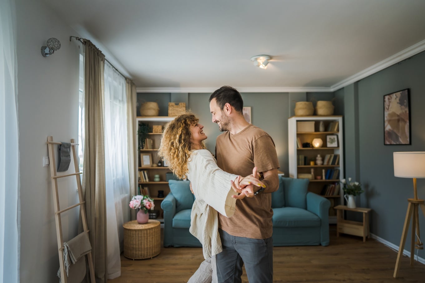 A man and a woman are dancing in the middle of their living room. In the background are bookshelves and furniture.