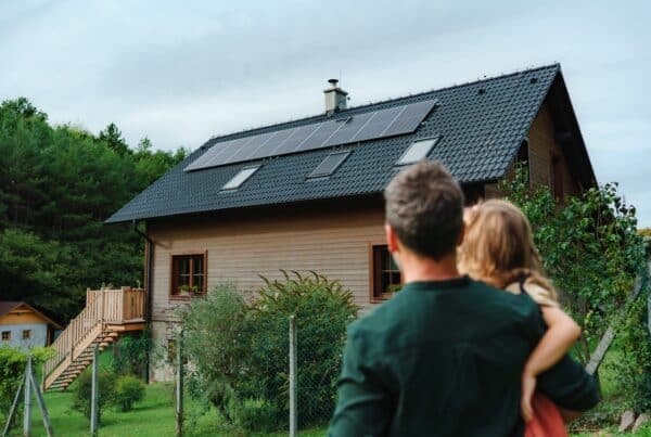 A man is holding a little girl in his arms. They are both looking at a house with solar panels.