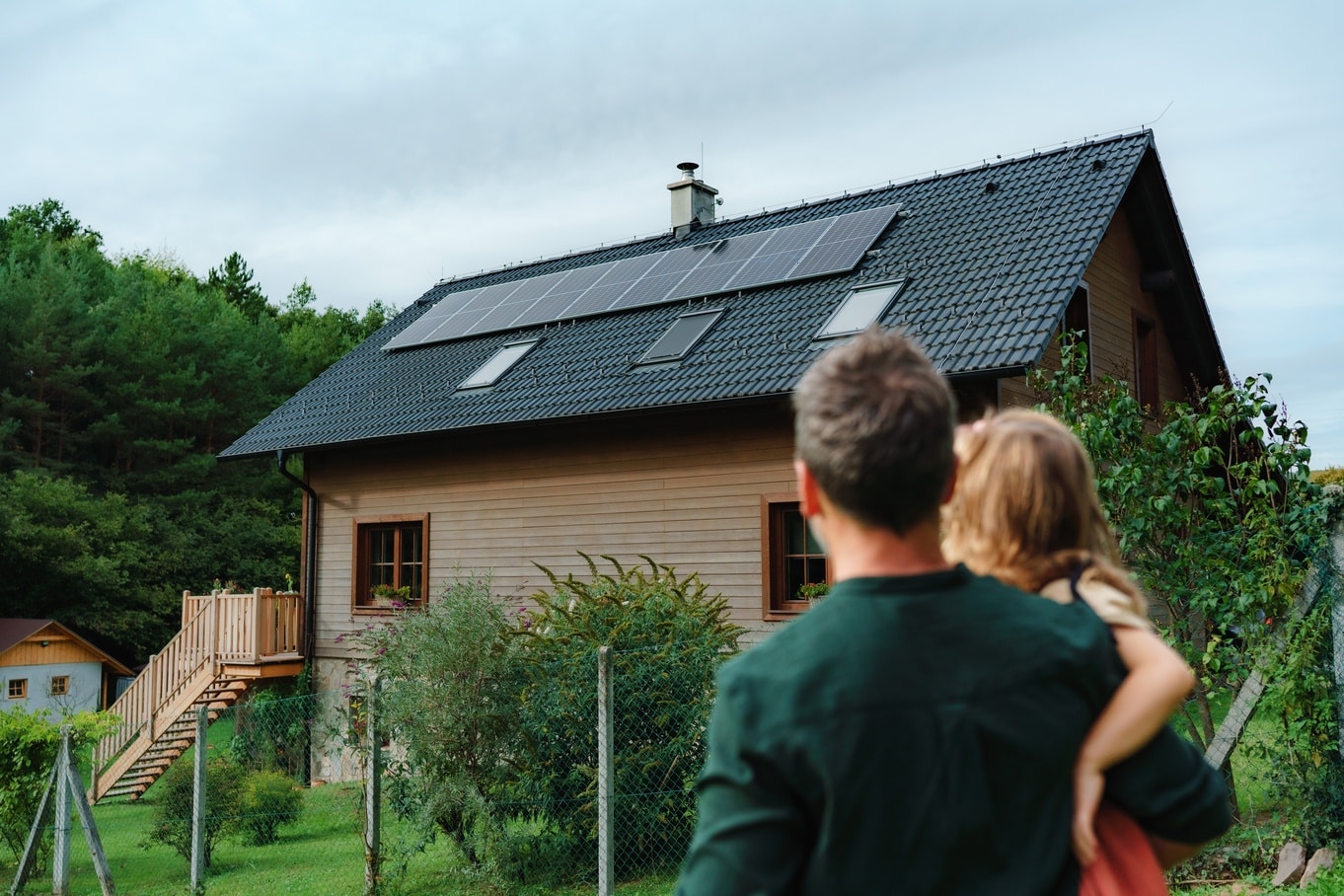A man is holding a little girl in his arms. They are both looking at a house with solar panels.