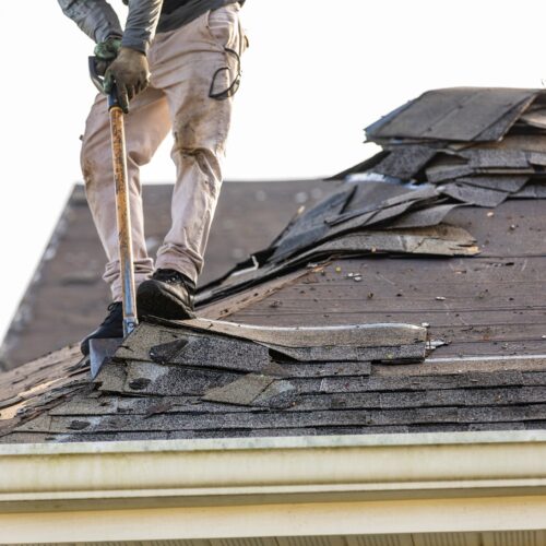 home improvement contractors A man stands on the roof of a house and removes shingles with a tool held by its handle, piling them up behind him.