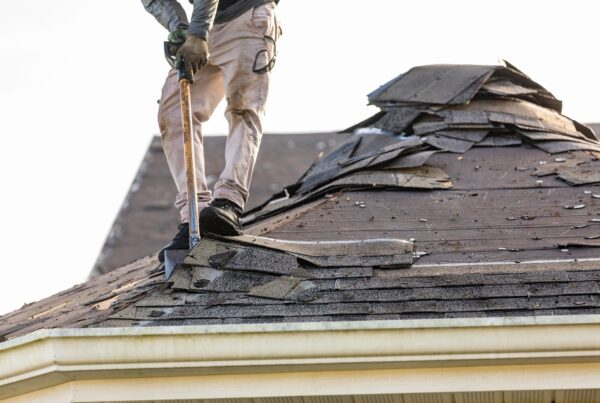 A man stands on the roof of a house and removes shingles with a tool held by its handle, piling them up behind him.