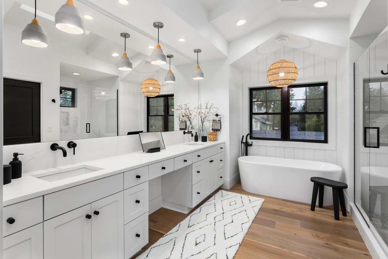 A beautiful, very white bathroom with a large window above the tub and a luxury vanity with a wall-length mirror.