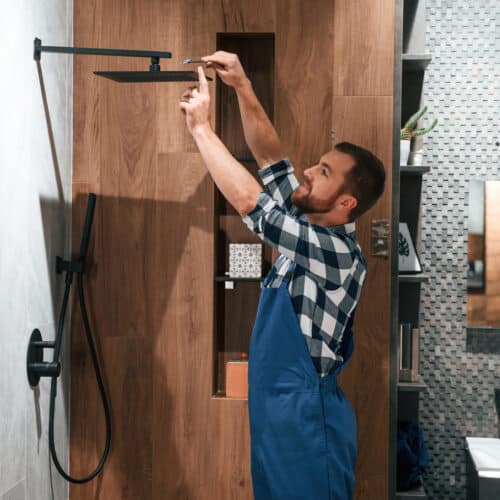 home improvement contractors A man with a beard, wearing overalls, is repairing a square, matte-black showerhead in a modern bathroom with wooden accents.