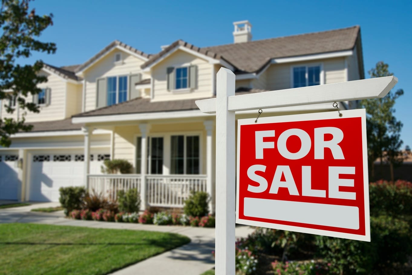 A large red “For Sale” sign is put up on the front lawn of a large residential home with beige siding and a dark-colored roof.