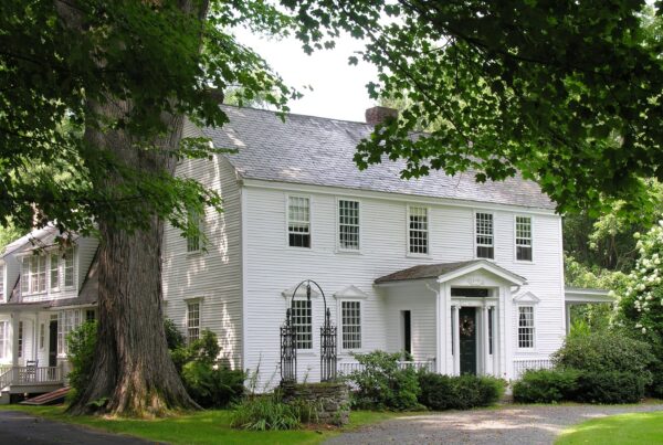 A white vintage home's exterior. Greenery surrounds the house with a large tree and stone well to the left.