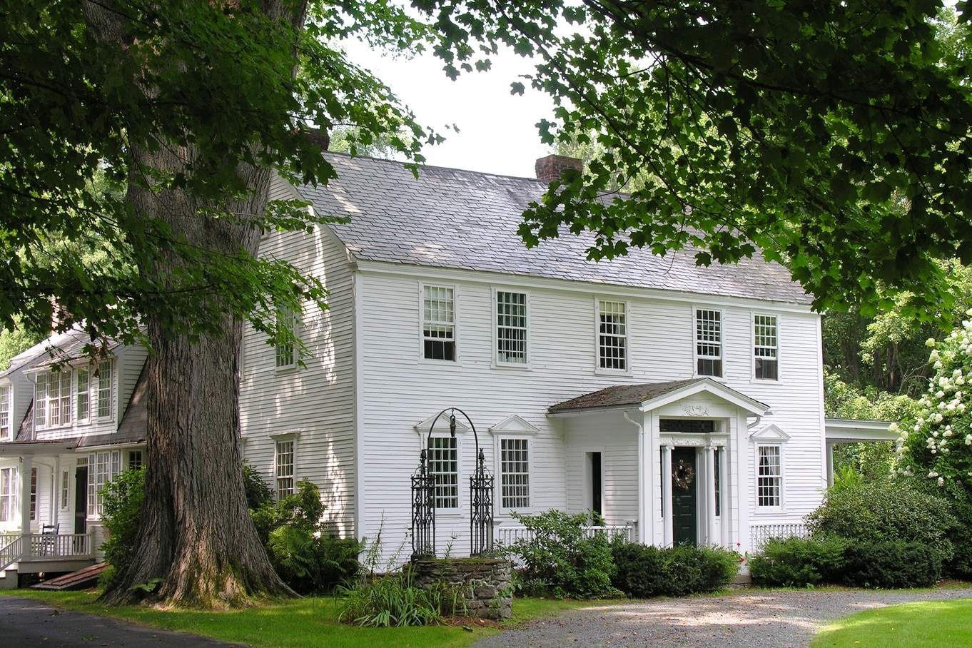 A white vintage home's exterior. Greenery surrounds the house with a large tree and stone well to the left.
