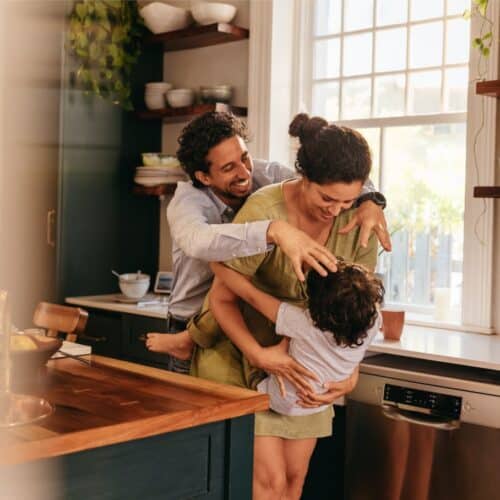 home improvement contractors Two young parents smile as they happily play with their child in the kitchen, between the sink and the island.