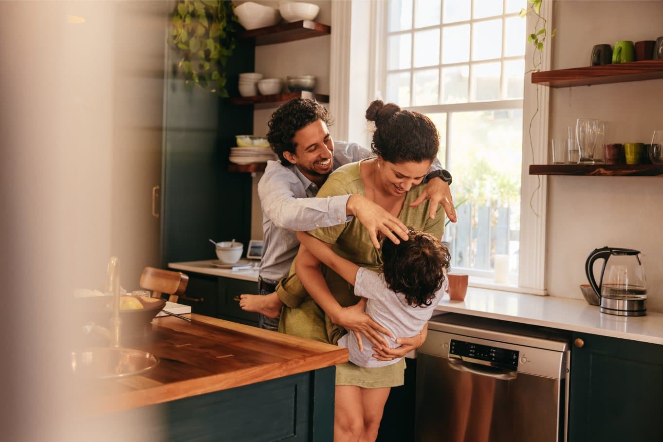 Two young parents smile as they happily play with their child in the kitchen, between the sink and the island.