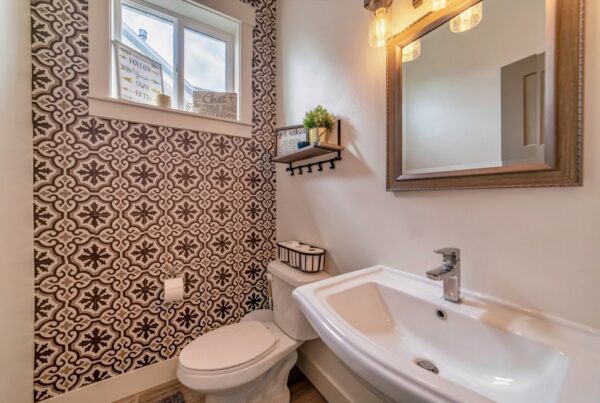 An interior view of a small bathroom with a toilet, sink, mirror, and a wall with patterned wallpaper.