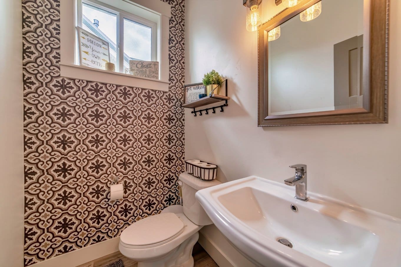 An interior view of a small bathroom with a toilet, sink, mirror, and a wall with patterned wallpaper.