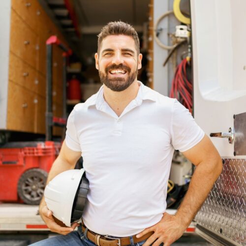home improvement contractors A smiling man wearing a white polo shirt has a hand on his hip and holds a white hardhat while standing in front of an open vehicle that has a variety of tools inside.
