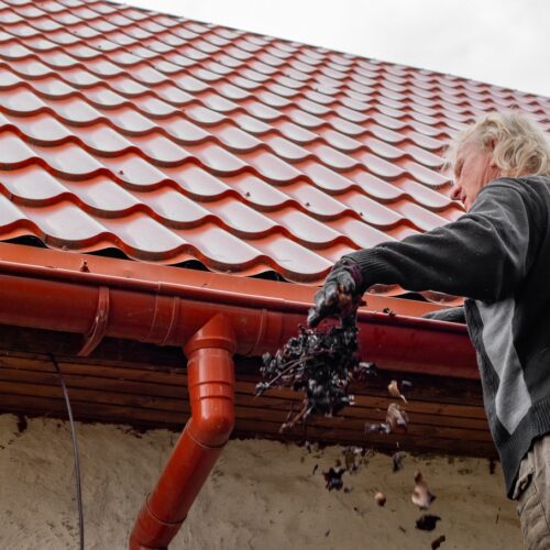 home improvement contractors A man stands next to the gutters on his red-tiled roof. He throws and handful of dead leaves down toward the ground.