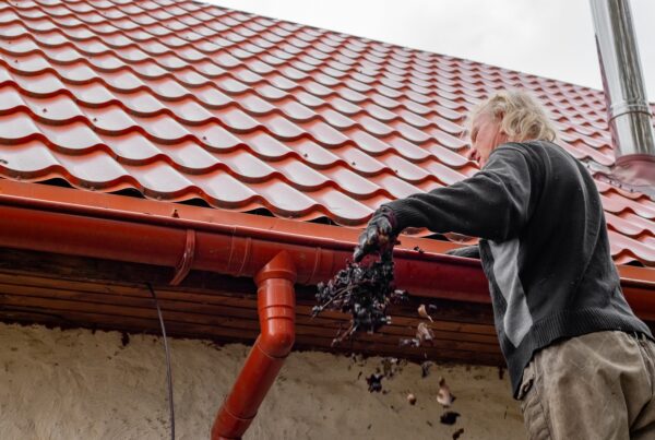 A man stands next to the gutters on his red-tiled roof. He throws and handful of dead leaves down toward the ground.