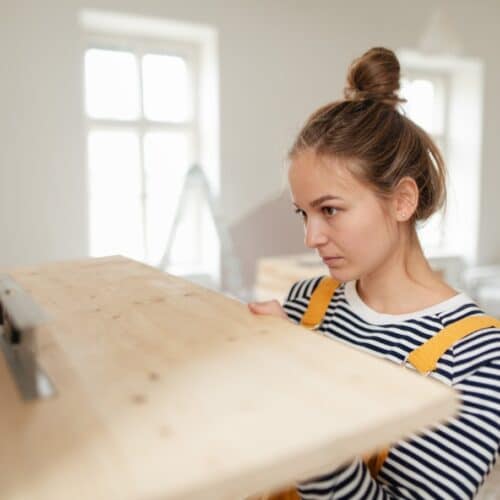 home improvement contractors A woman with her hair in a bun looks intently at a wooden shelf she is holding up to a wall. It has a level balanced on it.