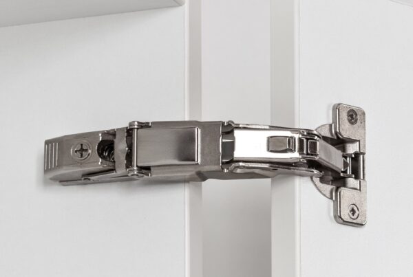 Close-up of a soft-close hinge on the inside of a cabinet and a cabinet door. The white cabinet door is open wide.