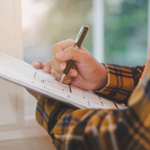 home improvement contractors A person wearing a flannel stands in front of a window and uses a pen to write on a clipboard containing house blueprints.