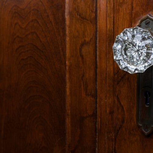 home improvement contractors A close-up of a glass doorknob with an ornate metal backplate on a dark wooden door with a visible grain.