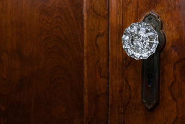 A close-up of a glass doorknob with an ornate metal backplate on a dark wooden door with a visible grain.