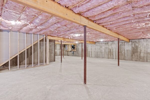 A wide view of an unfinished basement in a home with concrete floors and pink insulation in the ceiling.