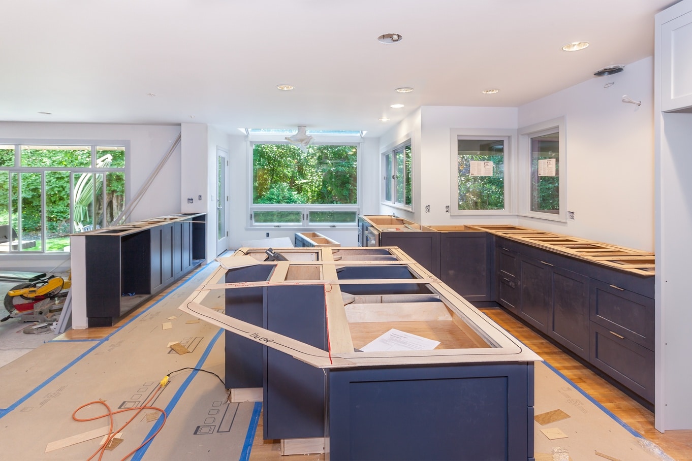 A kitchen renovation shows blue cabinets, large open windows, no countertops, and tools scattered across the floor.