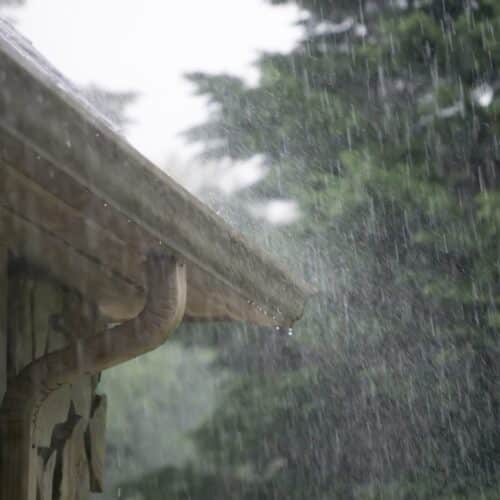 home improvement contractors Rain pours down and splashes off the side of a home's roof with pine trees in the background and a dark sky.