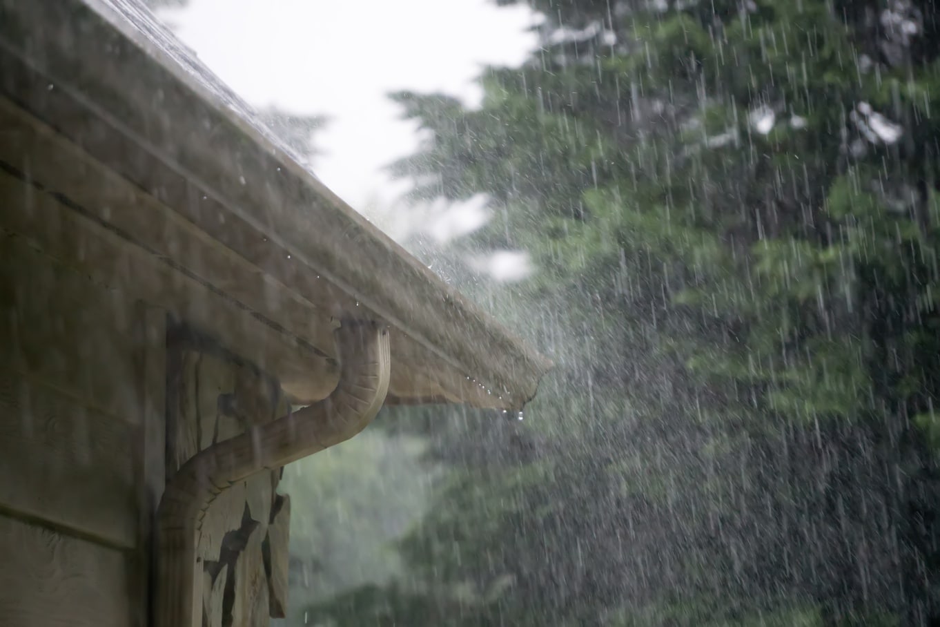 Rain pours down and splashes off the side of a home's roof with pine trees in the background and a dark sky.