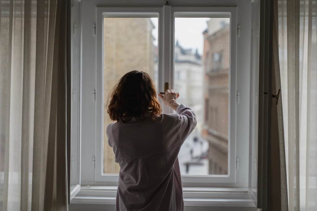 A person standing in front of a two-pane window and reaching up to twist the window's latch-style lock.