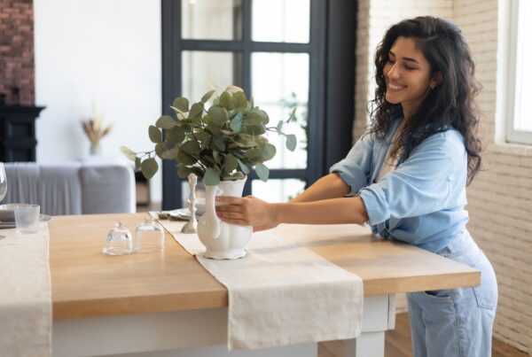 A smiling woman is placing a vase on a countertop. She’s wearing a blue shirt with the sleeves rolled up.