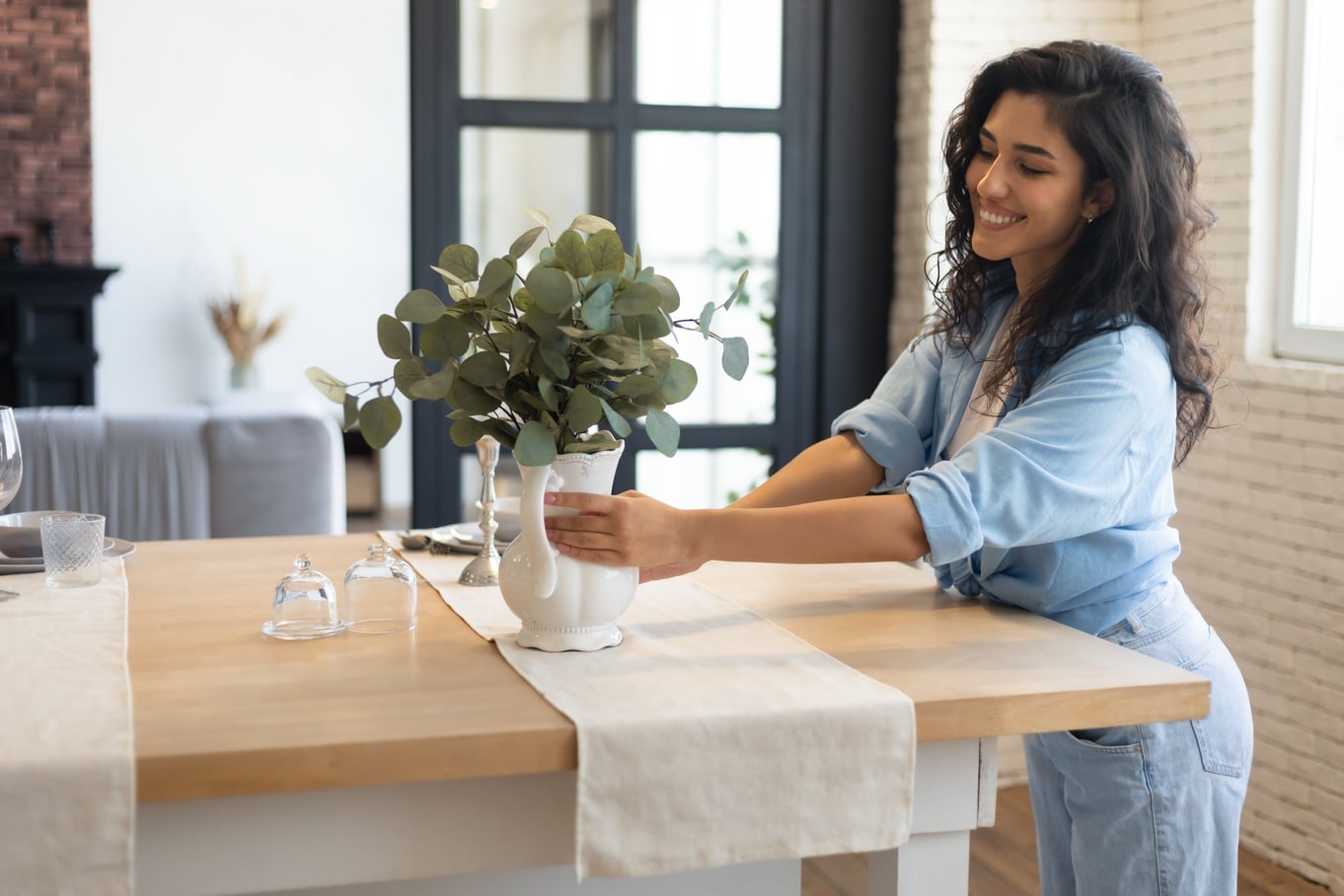 A smiling woman is placing a vase on a countertop. She’s wearing a blue shirt with the sleeves rolled up.