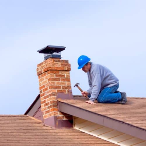 home improvement contractors A man in a blue hard hat kneels on a shingled roof, hammering beside a brick chimney with a black cap under a clear sky.