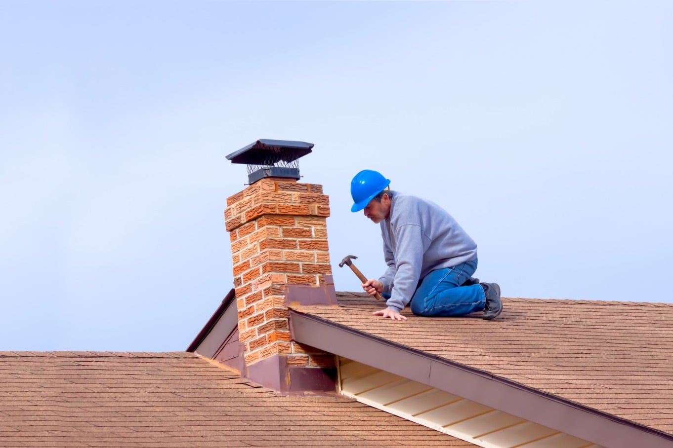 A man in a blue hard hat kneels on a shingled roof, hammering beside a brick chimney with a black cap under a clear sky.