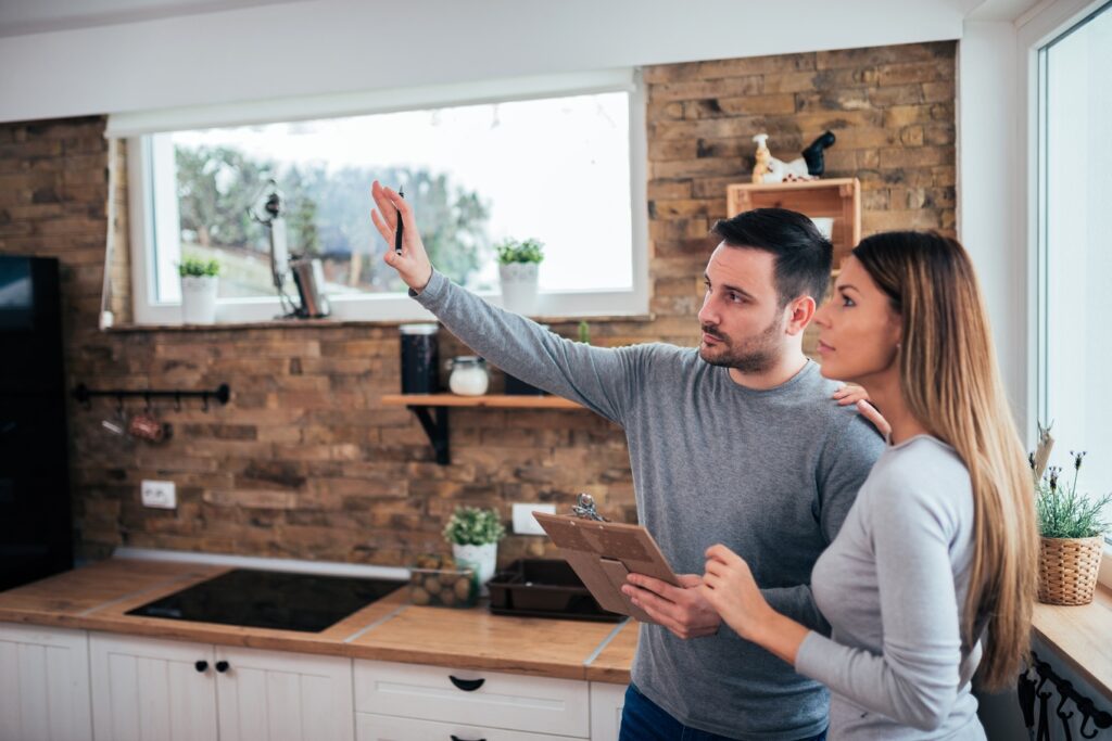 A man and woman stand in a kitchen with a stone accent wall. The man gestures at the room and holds a clipboard.