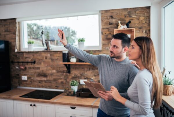 A man and woman stand in a kitchen with a stone accent wall. The man gestures at the room and holds a clipboard.
