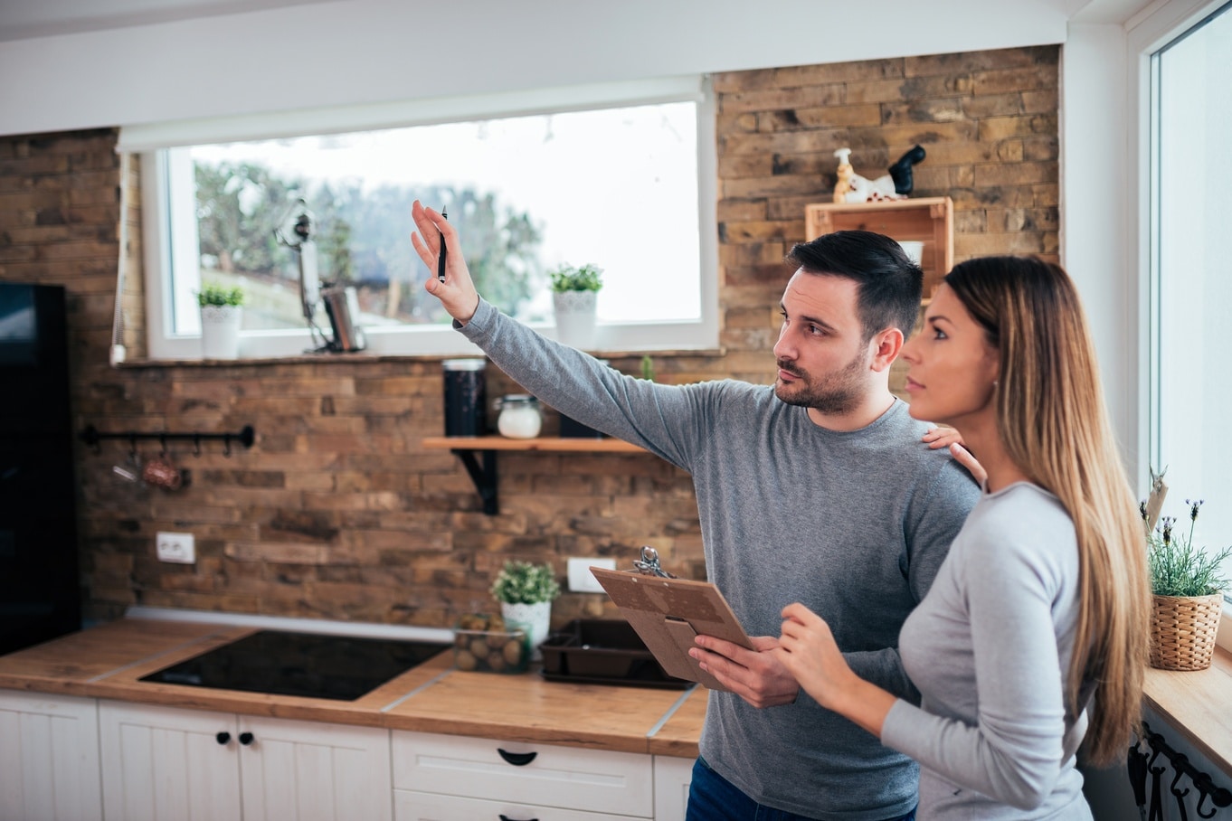 A man and woman stand in a kitchen with a stone accent wall. The man gestures at the room and holds a clipboard.