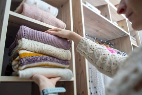 A woman placing a pile of folded up clothes into a cubby in her closet. Clothes are also hanging behind her.