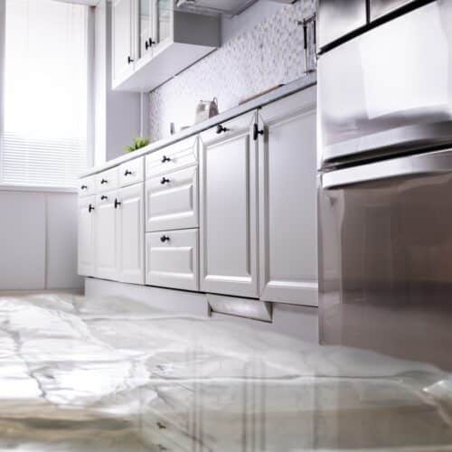 home improvement contractors A flooded kitchen with water covering the marble floor, white cabinets, and a stainless steel refrigerator.