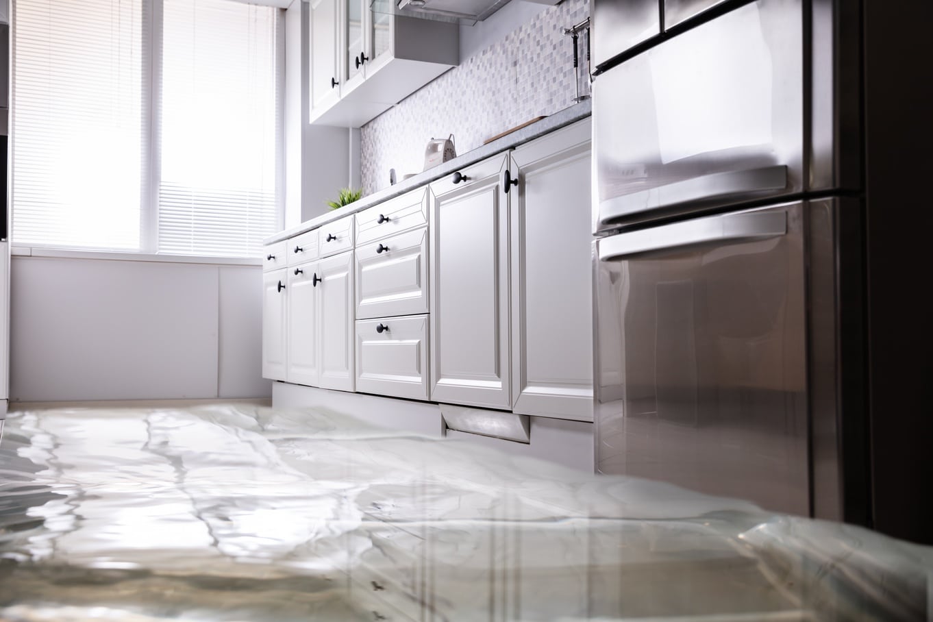 A flooded kitchen with water covering the marble floor, white cabinets, and a stainless steel refrigerator.