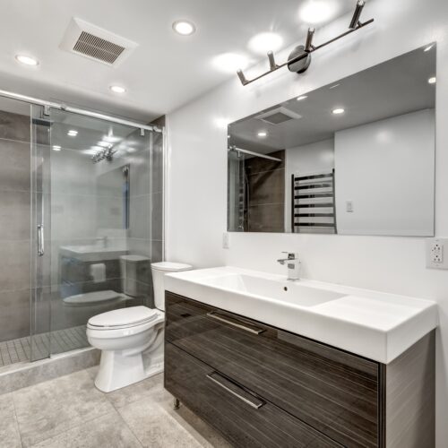 home improvement contractors An updated and modern bathroom features a walk-in shower with a sliding glass door, a toilet, and a large single-sink vanity.