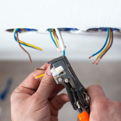 home improvement contractors The hands of an electrician using a wire stripper to strip the coating off of wires that are emerging from a white wall.