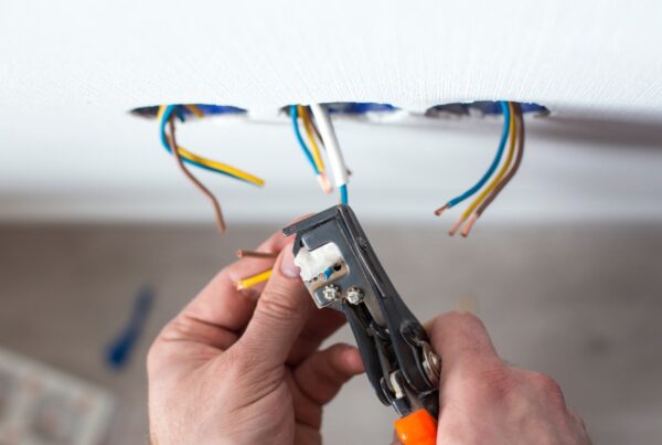The hands of an electrician using a wire stripper to strip the coating off of wires that are emerging from a white wall.