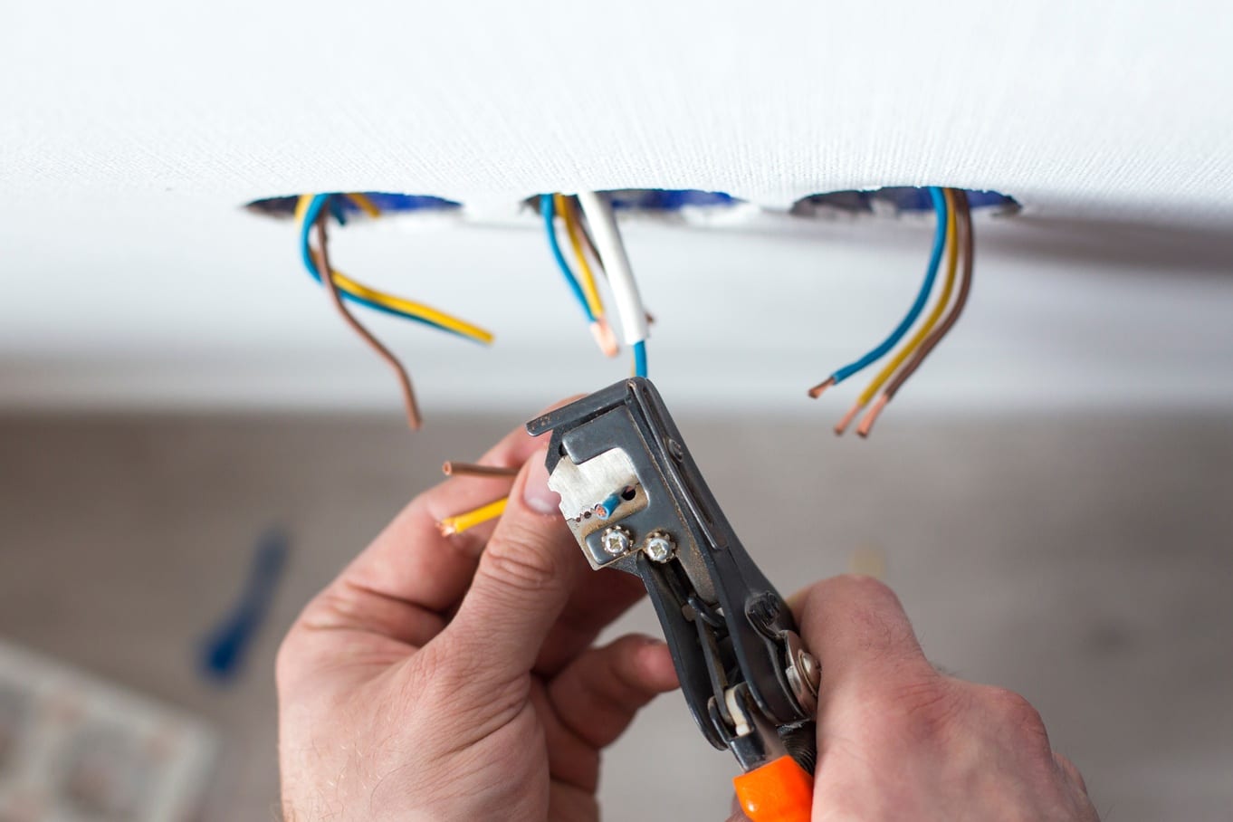 The hands of an electrician using a wire stripper to strip the coating off of wires that are emerging from a white wall.