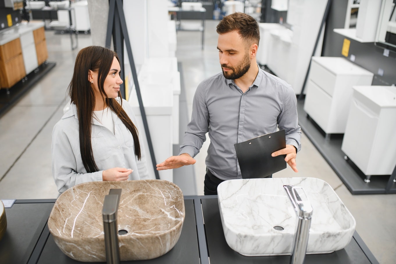 Two people standing in a showroom looking at bathroom sinks and faucets displayed on countertops inside a store.