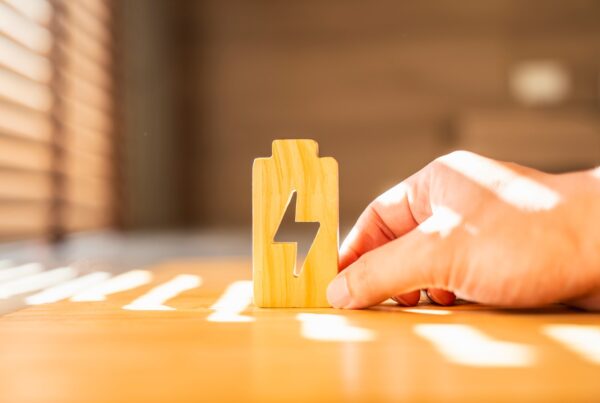 A person holding a wooden block on a wooden desk inside a home office. Sunlight shines inside through a nearby window.