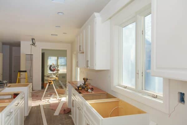 The interior of a home's kitchen during a remodel with white, unfinished kitchen cabinets, a tabletop saw, and more.