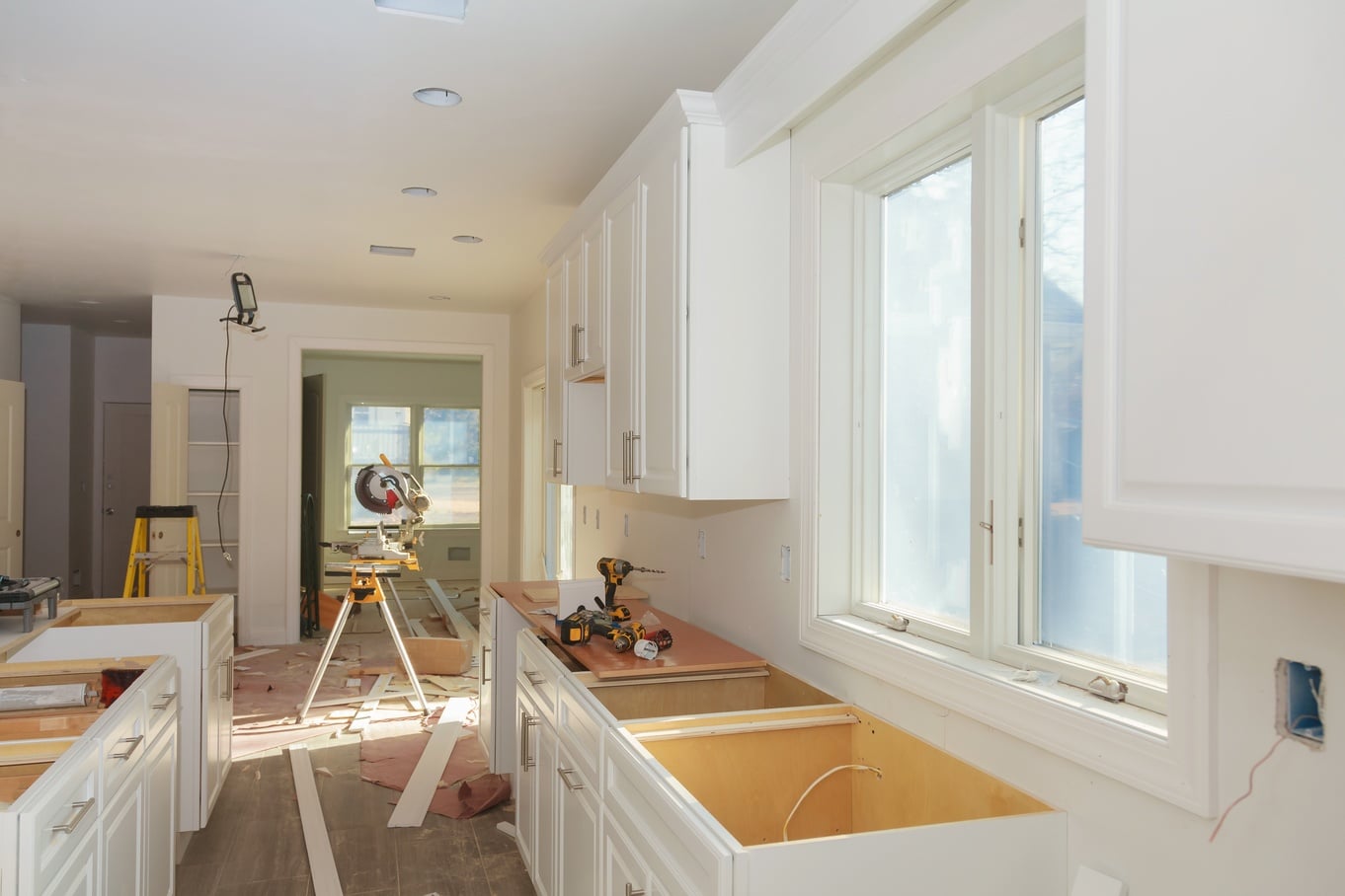 The interior of a home's kitchen during a remodel with white, unfinished kitchen cabinets, a tabletop saw, and more.