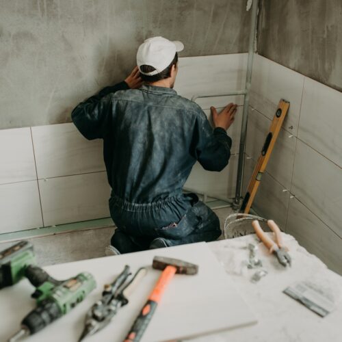 home improvement contractors A worker is kneeling and pressing a large white tile against the wall. Several construction tools are nearby.