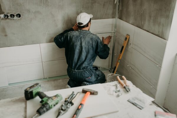 A worker is kneeling and pressing a large white tile against the wall. Several construction tools are nearby.