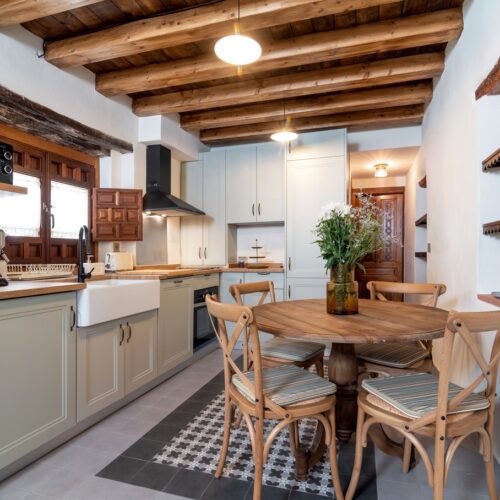 home improvement contractors Cozy farmhouse-style kitchen featuring a wood-beam ceiling, shaker cabinets, open shelving, and a round table with chairs.